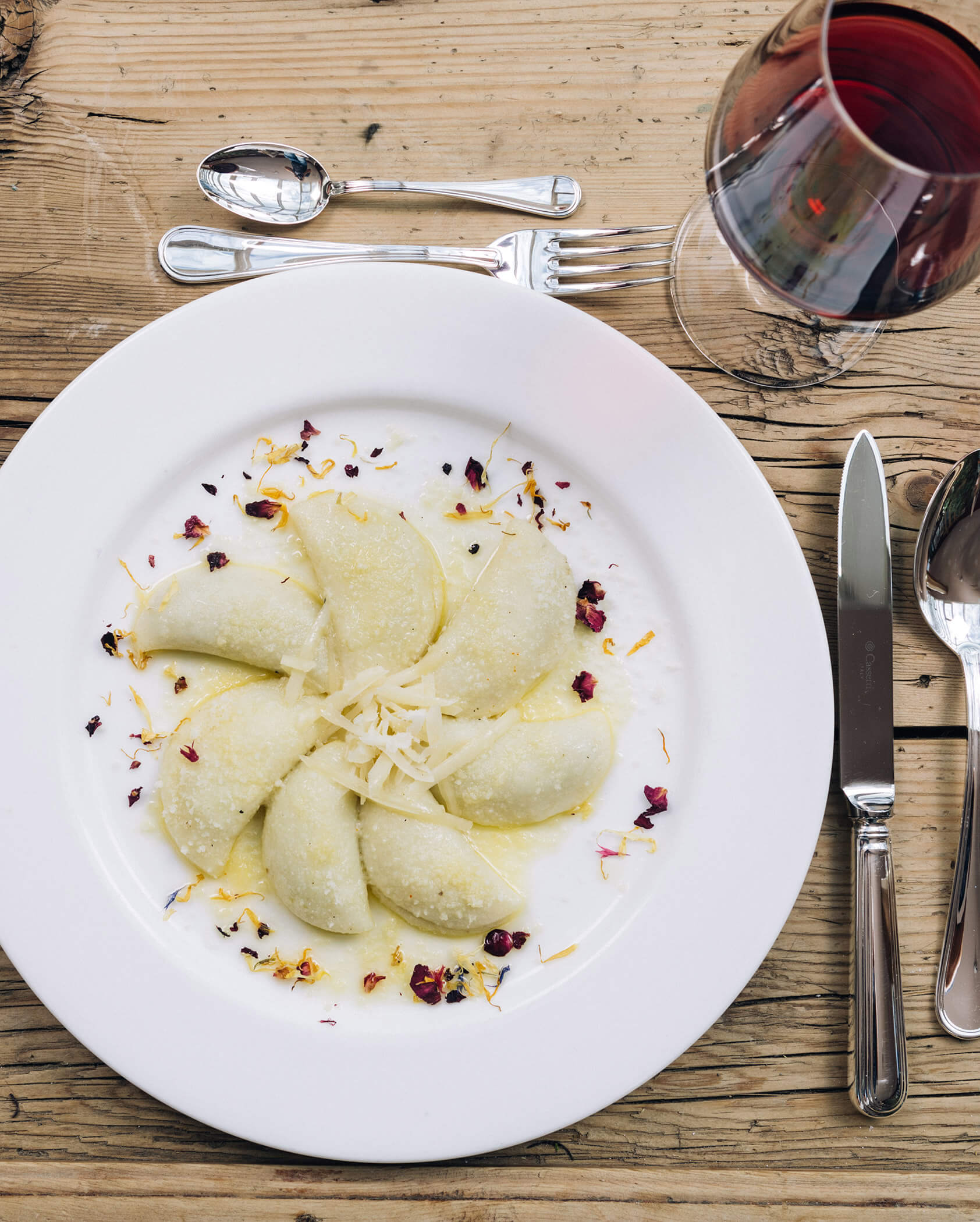 Un piatto di ravioli a mezzaluna con formaggio e petali di fiori, accanto a un bicchiere di vino rosso su un tavolo rustico. - Hotel Tenz