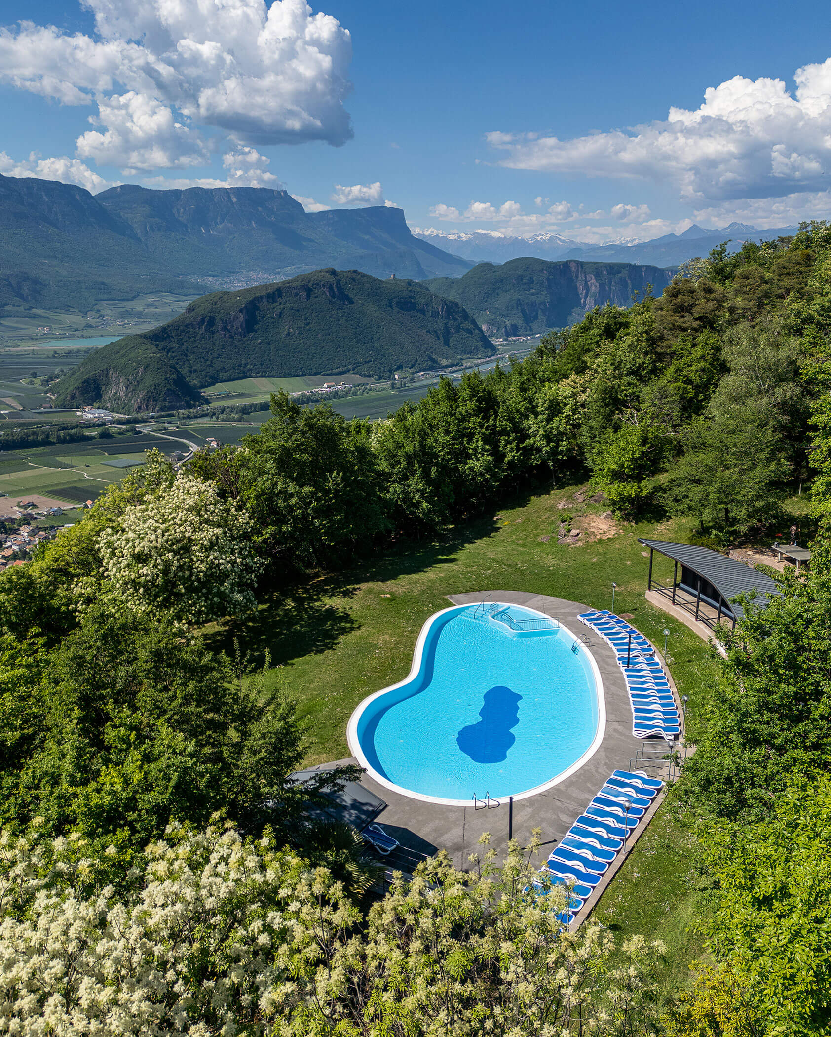 Ein herzförmiger Pool mit Liegestühlen, umgeben von Bäumen und Bergen unter einem teilweise bewölkten Himmel. - Hotel Tenz