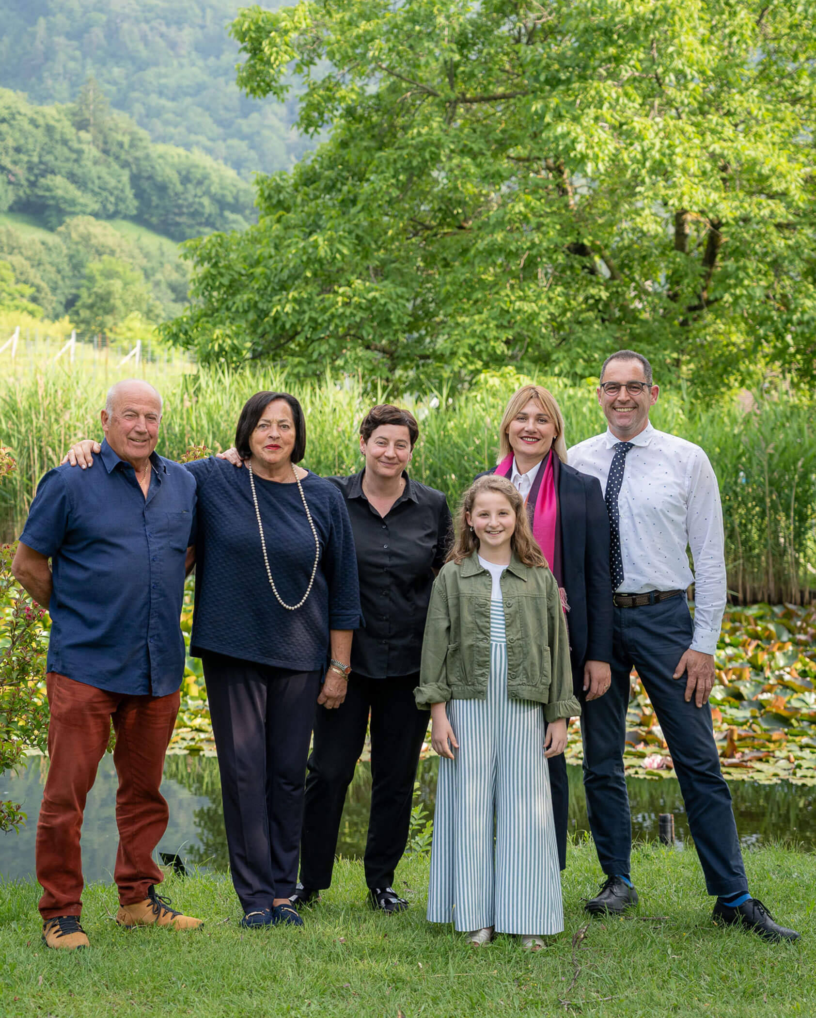 Sechs Personen, darunter ein Kind, stehen lächelnd im Freien vor Bäumen und einem Teich zusammen. - Hotel Tenz
