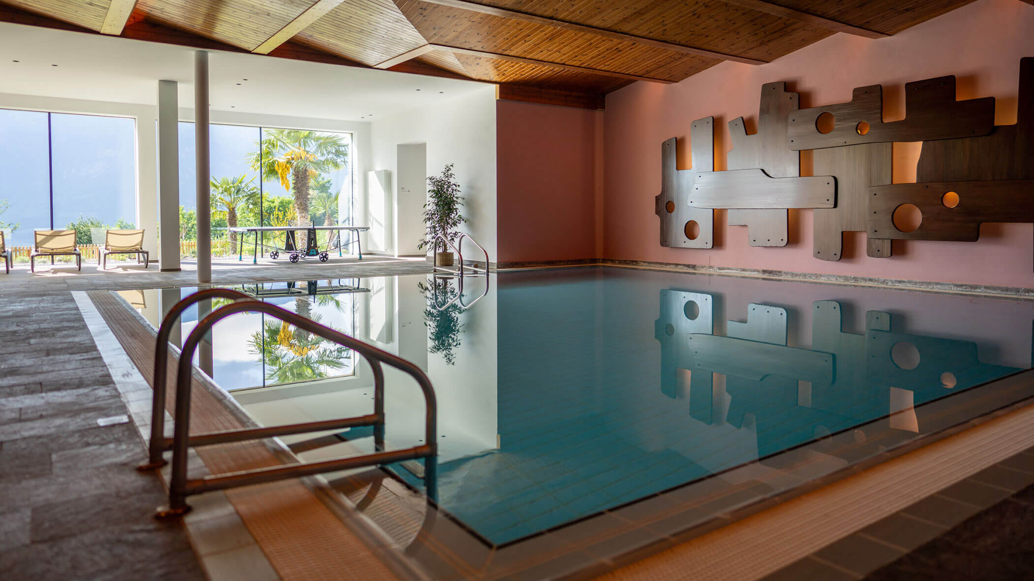 Innenpool mit moderner Wandkunst, großen Fenstern und Stühlen mit Blick auf einen Garten mit Palmen und Bergen. - Hotel Tenz